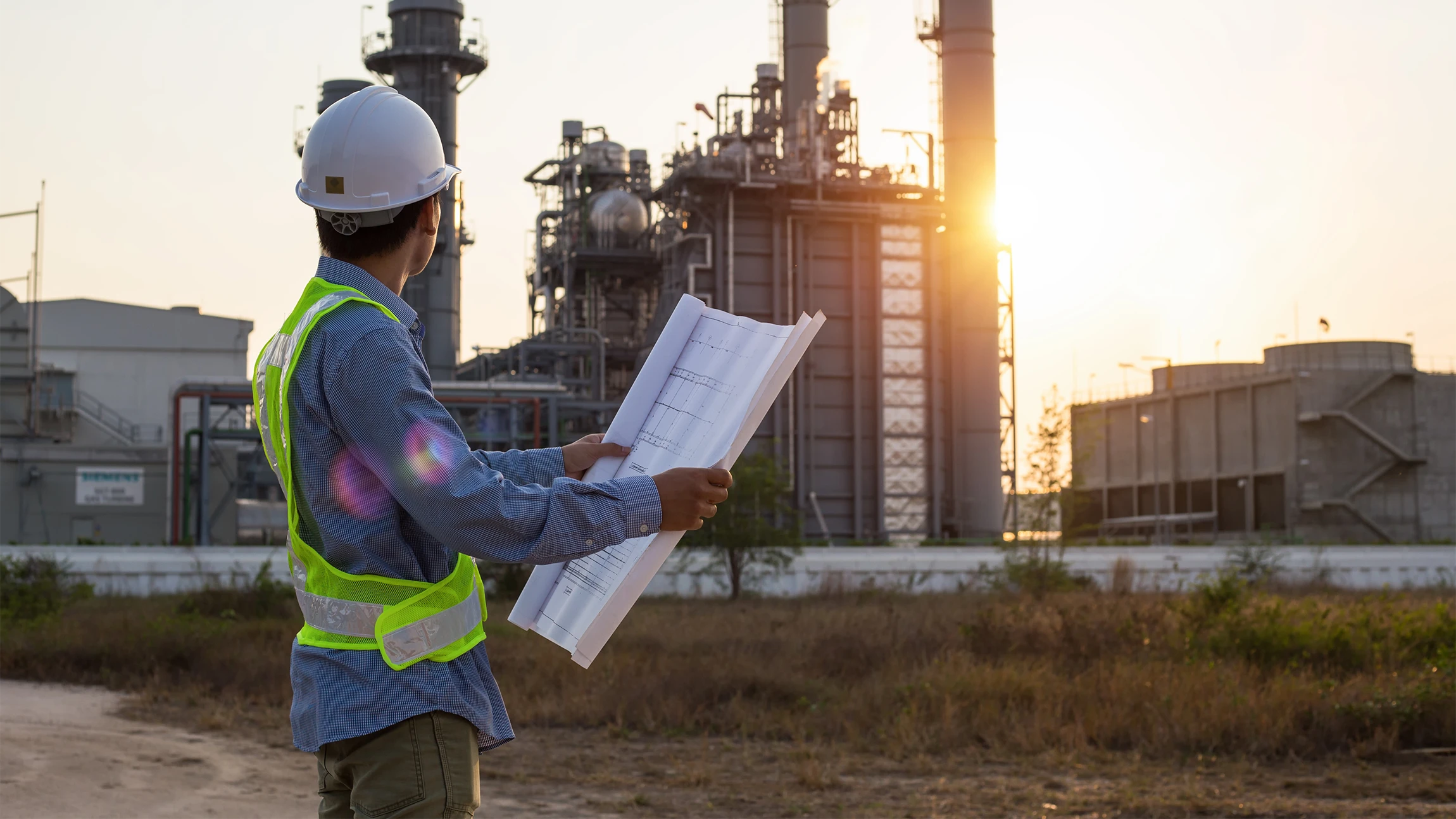 Engineer wearing a white hard hat and reflective vest holding blueprints and looking at an industrial plant at sunset.
