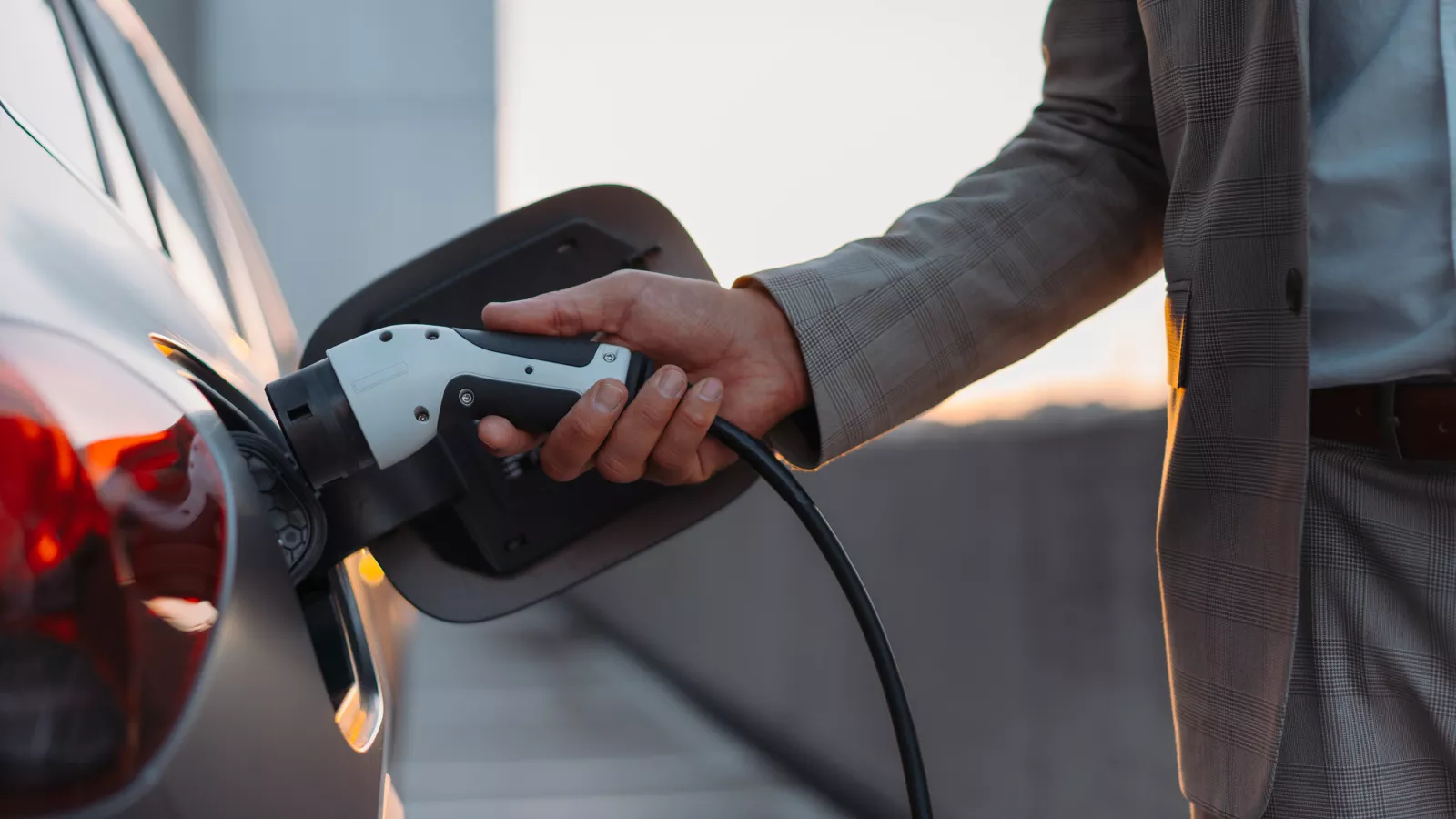 Ein Mann in einem eleganten karierten Anzug lädt sein Elektrofahrzeug an einer Ladestation auf. Der Fokus liegt auf seiner Hand, die das Ladekabel hält und mit dem Fahrzeug verbindet. Die Szene spielt sich auf einem Parkdeck bei Sonnenuntergang ab.