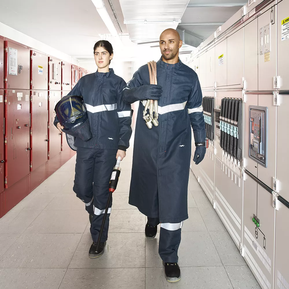 Two people in protective work uniforms walk through an industrial facility corridor, carrying tools and safety equipment.