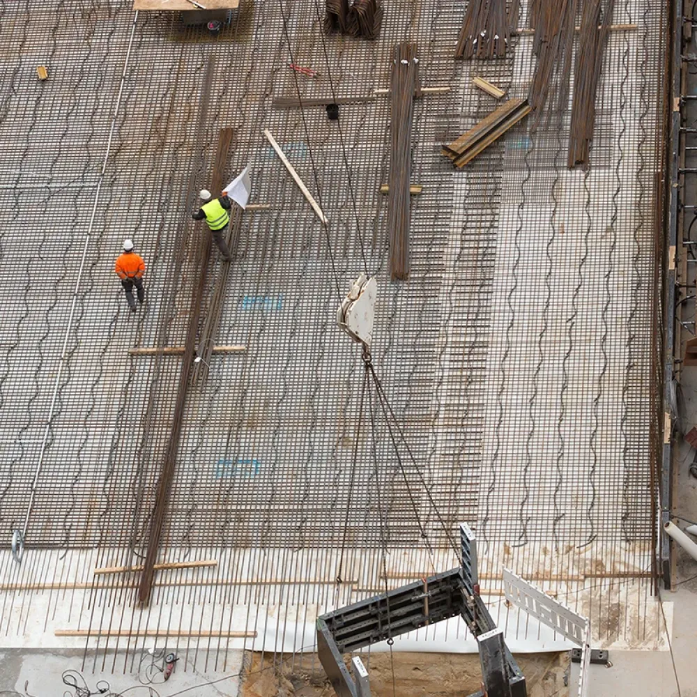 Overhead view of a construction site with two workers in safety helmets walking on a steel reinforcement grid with materials and equipment nearby.