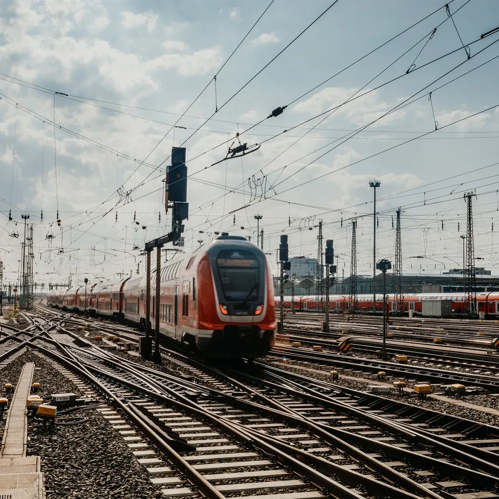 High-speed passenger train traveling through a rail yard with multiple tracks, overhead power lines, and signal systems under a cloudy sky.