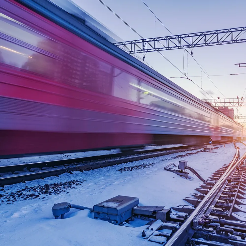 High-speed train passing a snow-covered railway junction at dusk, with blurred motion, overhead power lines, and visible switch components along the tracks.