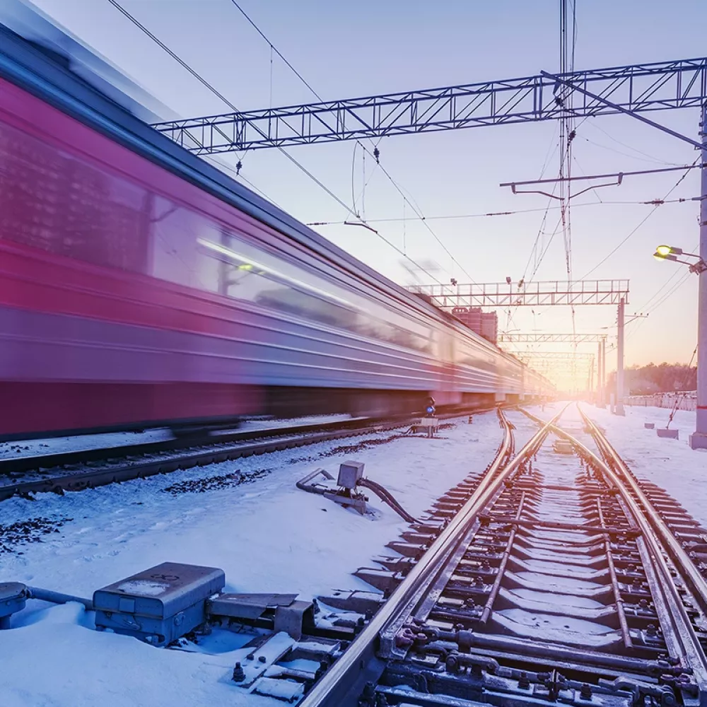 High-speed train passing a snow-covered railway junction at dusk, with blurred motion, overhead power lines, and visible switch components along the tracks.