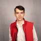 A striking image displays a young man in a vibrant red vest, his short, dark hair and friendly gaze standing out against a clean gray wall. The well-composed shot highlights the contrast of his white shirt and black zipper, making for an engaging and eye-catching portrait.