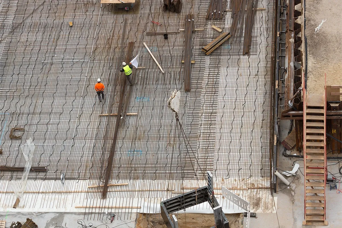 Overhead view of a construction site with two workers in safety helmets walking on a steel reinforcement grid with materials and equipment nearby.